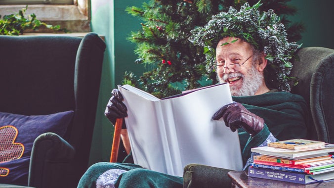 Green Father Christmas reads a story book while sitting in an armchair next to the Christmas Tree.
